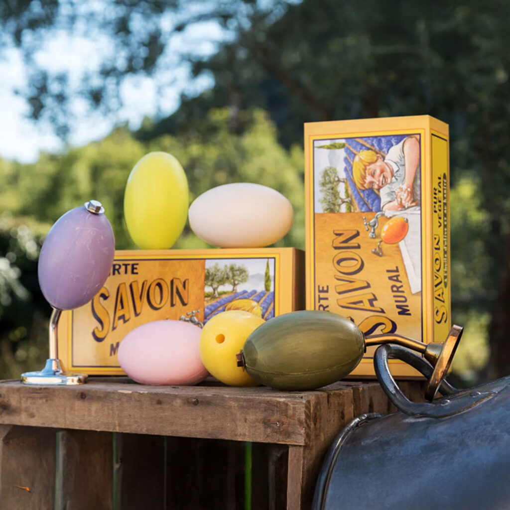 Collection complete des recharges de savons rotatifs de 260 grammes de La Savonnerie de Nyons et le coffret contenant un savon et le support mural en inox ou en laiton posés sur une cagette en boix vielli dans un decor provençal